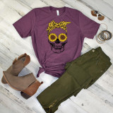Sunflower Skull FINAL STOCK Screen Print Heat Transfer, inside fashion ensemble with mauve t-shirt featuring a skull design with sunflower eyes and a yellow polka-dot bandana, paired with green cargo pants, tan ankle boots, and assorted jewelry on whitewashed wooden background.