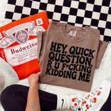 Hey Quick Question R U F*cking Kidding Me DTF Heat Transfer, humorous graphic design on beige T-shirt with black bold text, laid on a checkered black and white background, surrounded by a Budweiser poster, a person wearing white socks with red cherries and a graphic of a Converse shoe, and a space-themed hat.