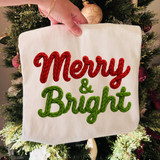 Faux Knit Merry & Bright DTF Heat Transfer, embroidered Christmas-themed decorative banner with red and green rope-style lettering, being held in front of a decorated Christmas tree.