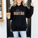 He's Hunting TAN DTF Heat Transfer product display on a black hoodie worn by a smiling woman holding iced coffee and making a peace sign.