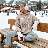 Cozy Girl Collage DTF Heat Transfer, fashion woman wearing beige hoodie with cozy girl design, sitting on wooden bench in snowy mountain town
