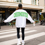 Oversized Cheermeister BUNDLE DTF Heat Transfer, front view of a person wearing a white oversized sweatshirt with green 'CHEERMEISTER' print on the back, walking across a crosswalk in an urban setting.