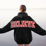 Oversized Believe BUNDLE DTF Heat Transfer, woman wearing a black hoodie with large red and white graphic text saying BELIEVE on the back, standing outdoors near water during sunset.