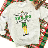 I Just Like To Smile Smiling Is My Favorite Screen Print Heat Transfer sweatshirt, featuring a festive design of a cheerful elf holding a sign with the quote, displayed on a plaid background with holiday decorations.