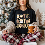 Tis The Season Leopard DTF Heat Transfer, Christmas-themed design on a black T-shirt with snowman, Santa, Christmas tree, and festive text, worn by a woman holding a marshmallow-filled mug with a Christmas backdrop.