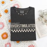 Probably Overstimulated CREAM DTF Heat Transfer, black T-shirt with white text and checkered pattern design displayed with crochet blanket, jewelry, and open book.