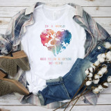 In A World Where You Can Be Anything Sublimation Transfer - Fashionable white t-shirt with a colorful global map heart design and inspirational quote, styled with casual denim jeans, plaid shirt, cotton flowers, and brown boots on a rustic wooden background.