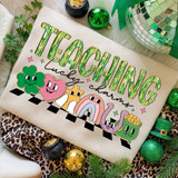 Teaching Lucky Charms Retro DTF Heat Transfer, colorful embroidered design of the word 'Teaching' with Lucky Charms characters like a four-leaf clover, heart, star, rainbow, cat, and pot of gold, used as a heat transfer for clothing or fabric decorations.