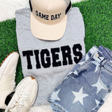 Tigers Fluffy Patch, front view of a starry design patch on denim shorts, beige baseball cap with 'GAME DAY' text, gray sweatshirt with 'TIGERS' in bold letters, white sneakers with brown accents, all arranged on green grass.