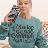 Make Sense Common Again DTF Heat Transfer, a woman holding a smartphone wearing a teal sweatshirt with the phrase 'Make Sense Common Again' printed on it.