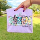 Patterned He Is Risen Easter DTF Heat Transfer, colorful Easter-themed design on a pastel purple clothing item.