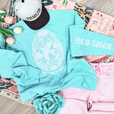 He Is Risen Fluffy Chenille Patch Set – 2 Piece Set, featuring a light blue sweatshirt with a white embroidered Jesus Christ design and text, and a matching light blue sleeve with embroidered words, placed on a colorful patterned rug with pink jeans, flowers, and a hat.