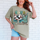 Howdy Easter Western Bunny DTF Heat Transfer, woman wearing a green t-shirt with Easter and western-themed design, including a cow print bow and leopard print bunny, against a white brick wall.