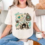 Western He Is Risen DTF Heat Transfer, illustration of religious symbols including a cave with a glowing light, a cross, a woman in traditional robes, and decorative elements on a white t-shirt worn by a woman sitting on a couch.