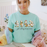Just A String Cheese Mama DTF Heat Transfer, humorous cartoon mama ducks with string cheese and coffee, shopping bags, and book, on a pastel blue t-shirt, in a cozy indoor setting