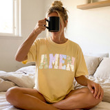 Faux Fabric Patchwork Amen DTF Heat Transfer, woman sitting cross-legged on bed holding a black mug with a yellow T-shirt with patchwork letter design.