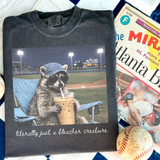 Literally Just A Bleacher Creature DTF Heat Transfer, humorous raccoon wearing sunglasses and a denim hat, sitting in a lawn chair and sipping iced coffee at a baseball stadium.