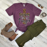 Living On A Prayer Leopard Print Screen Print Heat Transfer, image of a t-shirt with leopard print cross and the phrase 'Living On A Prayer' on the front, laid out on wooden floor with accessories and clothing nearby.