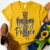 Living On A Prayer Leopard Print Screen Print Heat Transfer, yellow t-shirt with leopard print cross design and 'Living On A Prayer' text, surrounded by brown boots, blue jeans, sunflower bouquet, and hat.