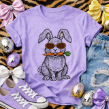 Cool Leopard Glasses Bunny DTF Heat Transfer, a purple t-shirt with a bunny graphic wearing leopard sunglasses and holding a carrot, surrounded by accessories like sneakers, bows, and decorative eggs.