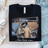 i Live Here Now Volleyball DTF Heat Transfer T-shirt, black t-shirt featuring a raccoon wearing sunglasses and a cap, sitting in a chair with a large iced coffee, with the slogan 'i live here now' at the bottom.