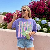 Lucky Duck Stripes DTF Heat Transfer, a colorful design featuring the word 'lucky' in green cursive and an illustration of a duck with striped wings on a purple T-shirt, worn by a smiling woman outdoors.