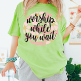 Worship While You Wait DTF Heat Transfer, a vibrant green t-shirt with a pink and black graphic design that features the phrase "worship while you wait" in stylish cursive font.