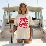 Salt Babe Stripes DTF Heat Transfer, a woman wearing a beige t-shirt with pink and red salt babe stripes DTF heat transfer design on front, standing on a boat.