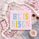 Floral Patterned He Is Risen DTF Heat Transfer, product image showing a pink cloth with the phrase 'HE IS RISEN' in colorful floral and plaid patterned letters, surrounded by decorative items such as sunglasses, jewelry, a drink, and a smartphone, on a soft, cozy background.