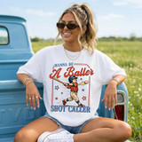 Wanna Be A Baller DTF Heat Transfer, a woman wearing a white t-shirt with baseball-themed graphic and text, sitting outdoors on a vintage truck in a field.