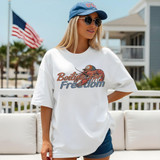 Boiling For Freedom DTF Heat Transfer, a woman wearing a white T-shirt with a lobster graphic and the text 'Boiling For Freedom', standing outdoors with American flags flying in the background.