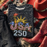 USA 250 Sequin Patch, patriotic embroidered patch with American flag colors, torch, and star design on a black textured T-shirt, surrounded by red cowboy boots, a red cowboy hat, and black distressed jeans on a checkered fabric background.