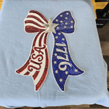 Close-up of oversized patriotic 1776 USA sequin bow patch heat pressed onto a light blue crewneck sweatshirt with rhinestone and glitter detailing.