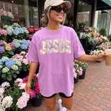 Faux Pearl Jesus DTF Heat Transfer, image of a woman wearing a pink T-shirt with 'JESUS' written in decorative faux pearl design, standing outdoors with colorful flowers in the background.