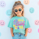 Catch Me at the County Fair DTF Heat Transfer, girl wearing a light blue T-shirt with colorful fair-themed design and text, standing in front of a wall with smiley face patterns.