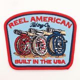 Reel American Fishing Co. Hat Patch 4 x 3 inches, embroidered patch with fishing reels and fishing rods design, featuring red, blue, white, and black colors, with text "Reel American" and "Built in the USA".