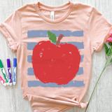 Apple Blue Stripes DTF Heat Transfer, pink T-shirt with a large red apple and blue stripes design featuring green leaf and brown stem, laid flat on a light wooden surface with colorful markers and pink flowers around it.