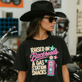 Raised On Backroads & Gas Station Snacks DTF Heat Transfer, a woman wearing a black T-shirt with a pink and teal retro gas pump graphic and colorful text in an indoor setting.