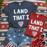 Land That I Love Fluffy Patch, patriotic T-shirts with American flag heart designs, denim shorts with star prints, and red Adidas sneakers on a star-spangled blanket.