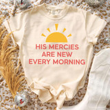 His Mercies Are New Every Morning DTF Heat Transfer, a design printed on a cream-colored t-shirt featuring a yellow sunrise graphic with pink text.