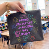 Helping Little Humans Understand Their Feelings DTF Heat Transfer, colorful design on a dark fabric tote bag featuring icons of hearts, speech bubbles, leaves, puzzle pieces, and hands.