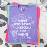 Helping Little Humans Understand Their Feelings WHITE DTF Heat Transfer, close-up of a purple T-shirt with white printed design and text, folded on a pile of colorful shirts.