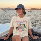 Meet Me At The Lake DTF Heat Transfer, a person sitting on a boat wearing a cream T-shirt with colorful laundry and lake scenery illustration, at sunset with boats on the lake in the background.