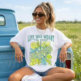 Life Was Meant For Great Adventure DTF Heat Transfer, a woman wearing a white T-shirt with a colorful adventure-themed design, sitting outdoors by a blue vehicle.
