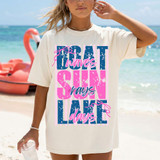 Boat Waves Sun Rays Lake Days DTF Heat Transfer, woman wearing a T-shirt with a colorful beach-themed graphic design, standing by the lake with a paddle boat in the background.