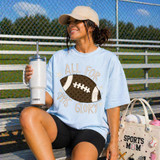 All For His Glory Football DTF Heat Transfer, a woman sitting on bleachers at a sports field wearing a light blue t-shirt with a football graphic and the phrase 'All For His Glory' printed on it.