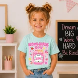 Is My Jam Back to School DTF Heat Transfer for Grades Pre-K–4, image of a young girl wearing a light blue T-shirt with colorful classroom-themed design and text, smiling indoors.