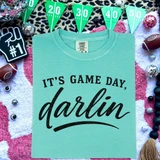 It's Game Day, Darlin' DTF Heat Transfer, turquoise T-shirt with black text, surrounded by football-themed decorations.