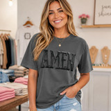 Amen Black Metallic Puff Heat Transfer, woman wearing a dark grey t-shirt with black puffed metallic letters spelling 'AMEN' in a boutique clothing store.