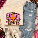 Pink Glitter Mascot - 62 teams - DTF Heat Transfer, a colorful graphic t-shirt with a lightning bolt and stars design, laid out on a cozy surface with denim jeans, sneakers, and accessories around it.